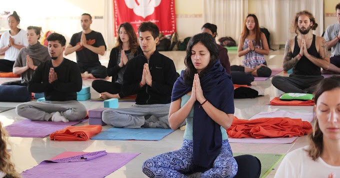 A group of people meditate on yoga mats in a brightly lit room. They sit cross-legged with hands in prayer position, eyes closed. A red and white banner hangs on the wall.